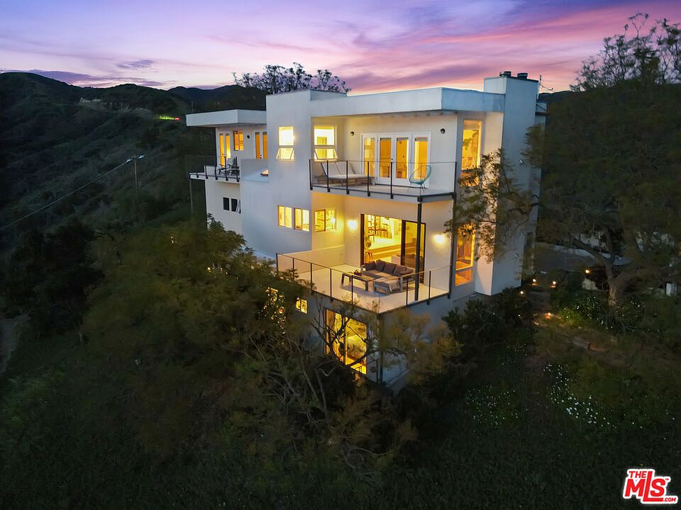 The home seen from the back at twilight, with warm interior light glowing through walls of glass and wraparound balconies cantilevered over the canyon