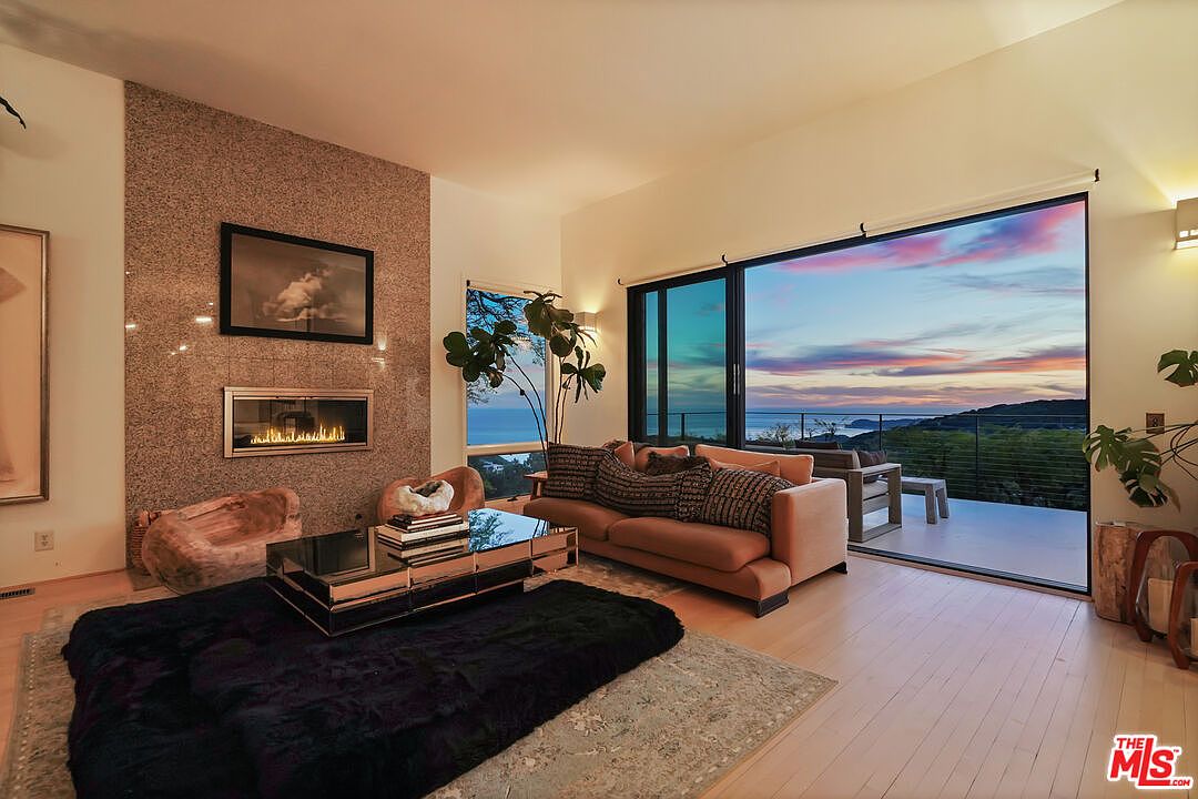 Living room with wood-burning fireplace, Fleetwood pocket doors fully open to a wraparound balcony framing the Pacific Ocean at twilight
