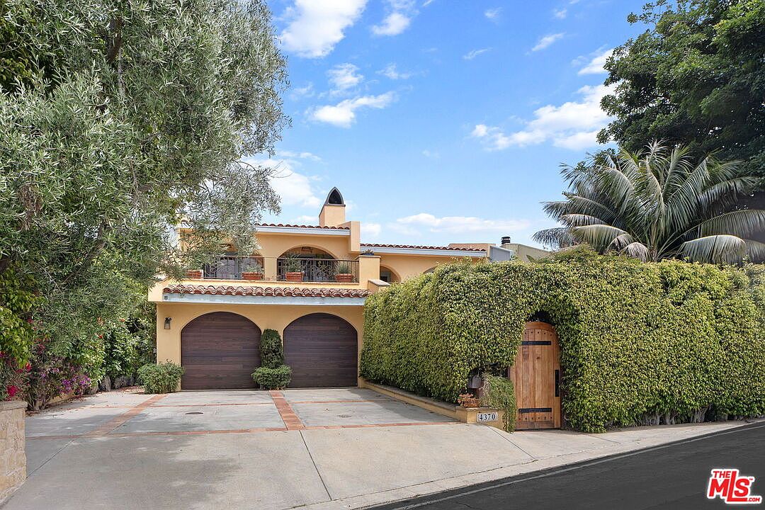 Spanish-style facade in warm ochre with a terracotta tile roof, hedge-walled entry, and twin garage doors